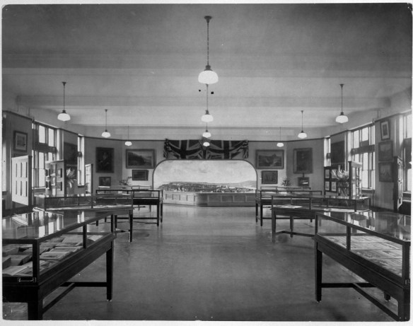 Photo noir et blanc d’une grande salle présentant des tables d’exposition de part et d’autre et des luminaires suspendus au plafond. À l’arrière-plan, on aperçoit une grande maquette de ville.