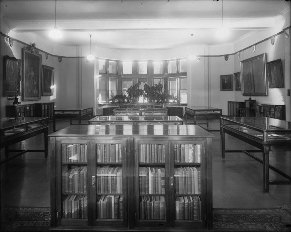 Photo noir et blanc d’une salle où se trouvent des bibliothèques et des vitrines d’exposition. À l’arrière-plan, on voit des fenêtres et des plantes.