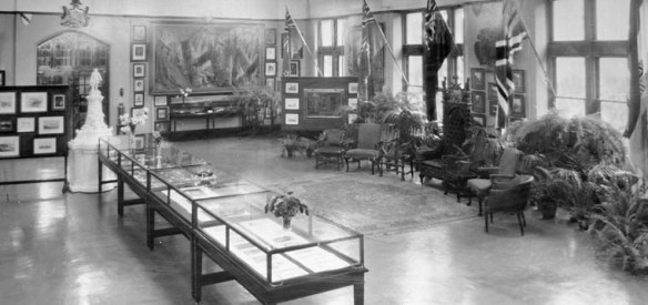 Photo noir et blanc d’une grande salle où se trouvent des vitrines d’exposition, une statue, des drapeaux, des plantes, des images encadrées, des chaises et un trône.