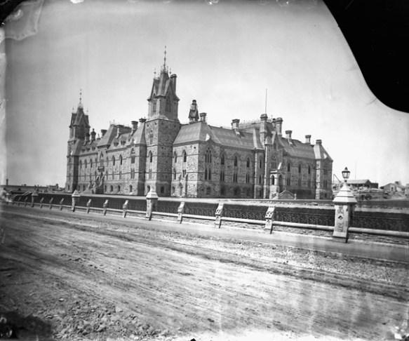 Photo d’un imposant édifice de pierre surmonté de tours. Devant, on voit un chemin de terre, bordé d’un muret en pierre et en fer forgé.