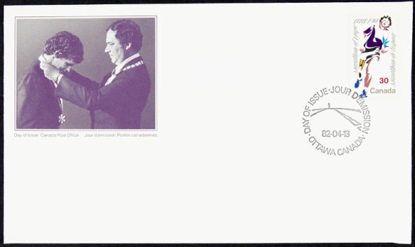 Un rectangle dans lequel figure la photo d’un homme remettant une médaille enrubannée à un autre homme, dont la tête est penchée vers l’avant. Dans le coin supérieur droit figure un timbre tamponné comprenant le dessin d’un homme en train de courir ainsi qu’un cachet de la poste mentionnant « Day of issue, Jour d’émission, Ottawa Canada, 82-04-13. »