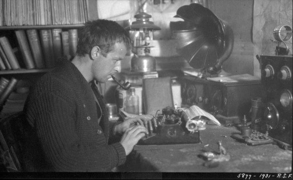 Un homme vêtu d’un tricot et fumant la pipe est assis à un bureau. Autour de lui se trouvent une bibliothèque, une lanterne et une radio.