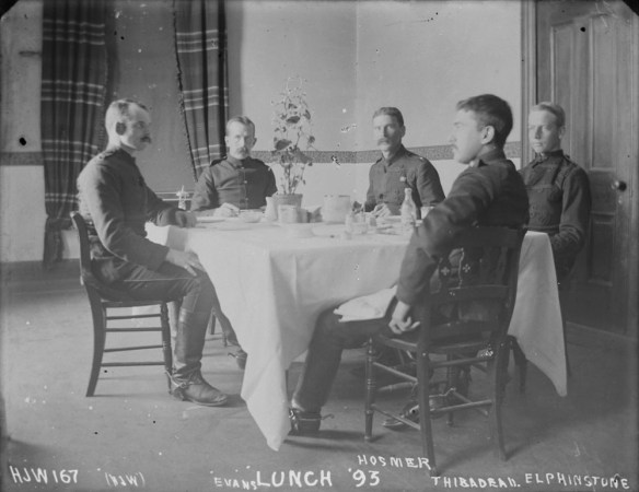 Cinq hommes autour d’une table couverte d’une nappe blanche. Une plante se trouve au centre de la table, sur laquelle sont aussi posées des bouteilles, des assiettes et de la vaisselle.
