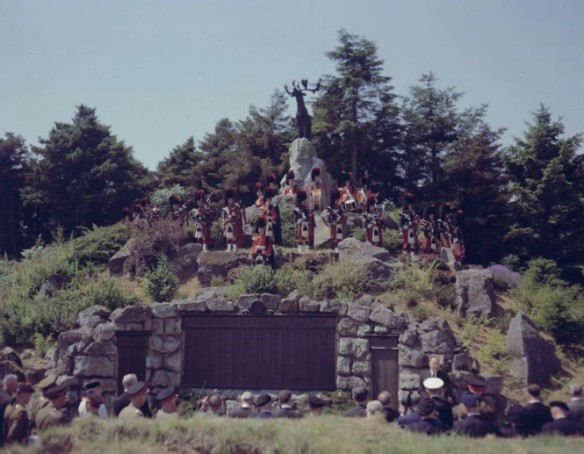 Groupe de soldats debout sur une colline, devant un monument représentant un caribou. Au pied de la colline, une foule devant des plaques de bronze observe la scène.