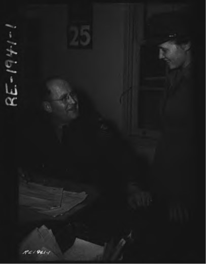 Un homme en uniforme (au centre de la photo), assis et portant des lunettes, regarde une femme en uniforme debout (en bordure droite de la photo).