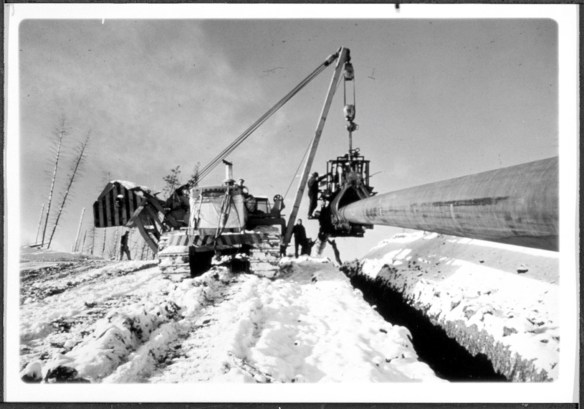 Machine Caterpillar avec système de levage et grappin soulevant un pipeline directement au-dessus du fossé où il sera déposé. Le fossé est bordé de neige. Plusieurs ouvriers se tiennent dans la neige et un autre est debout sur le grappin. Deux grands arbres, du côté gauche de l’image, ont des branches courtes sans feuilles.
