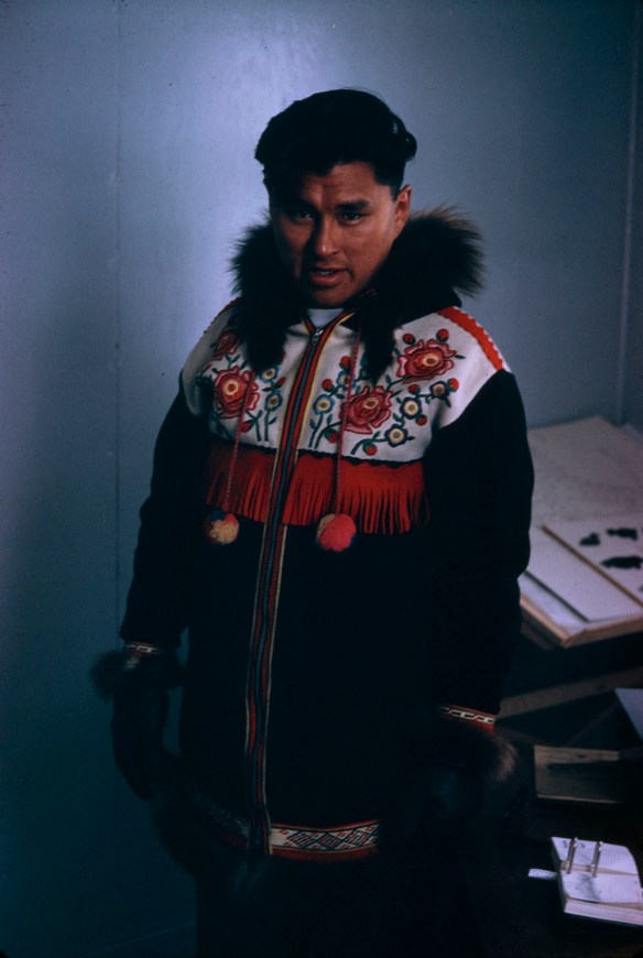 Photographie en couleur d’Abe Okpik, debout à l’intérieur, et vêtu d’un parka inuit noir avec broderie florale rouge, jaune et vert sur un empiècement blanc avec frange rouge. Il porte des gants noirs en fourrure. 