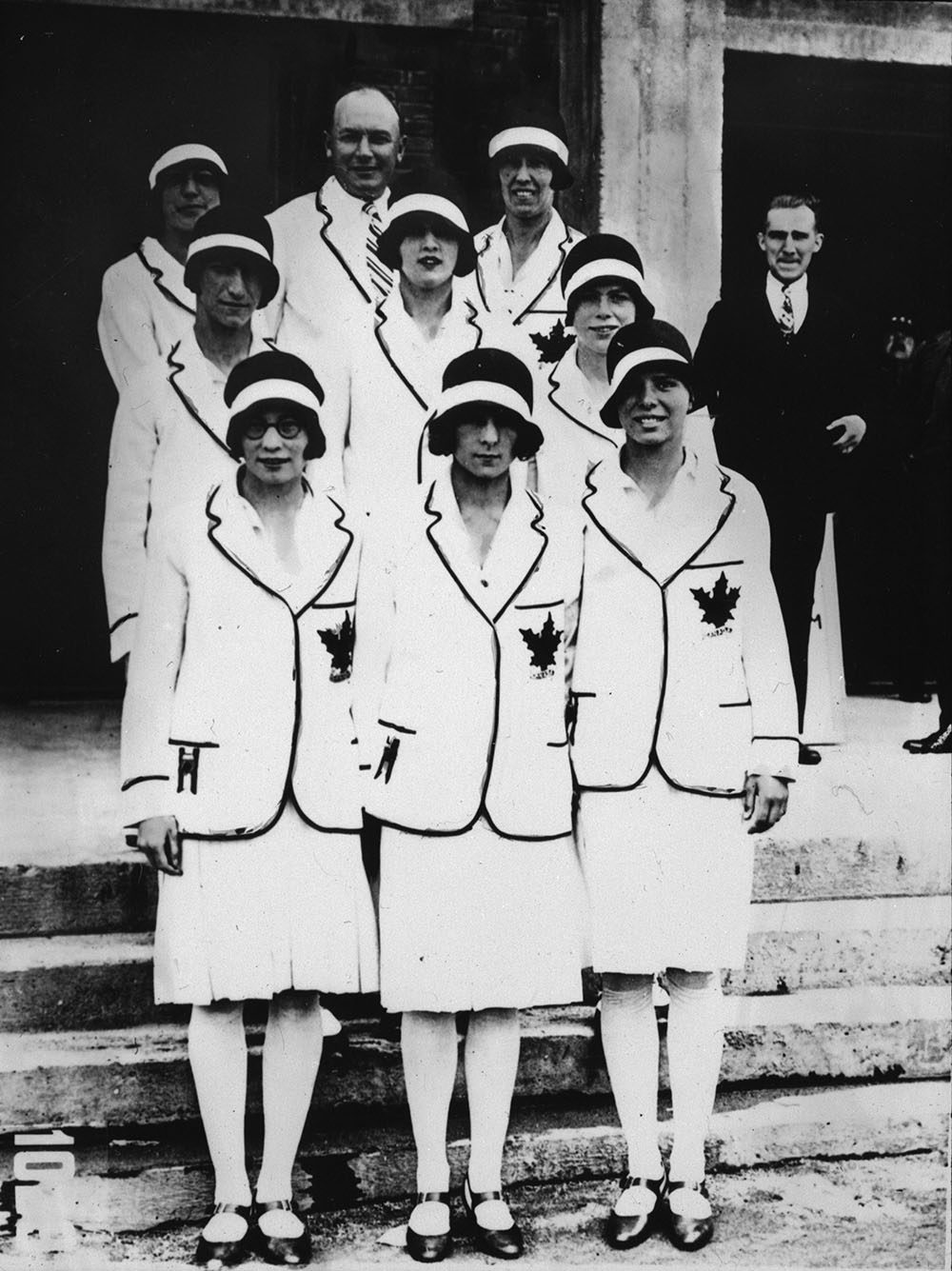 Photographie montrant six femmes vêtues d’une veste et d’une jupe blanches. On peut voir quatre autres personnes à l’arrière-plan.