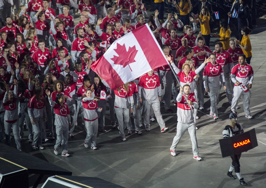 Photographie de nombreux athlètes marchant derrière un homme qui agite un grand drapeau du Canada.