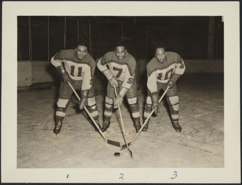 Photographie en noir et blanc de trois joueurs de hockey noirs alignés, leurs bâtons posés sur la glace.